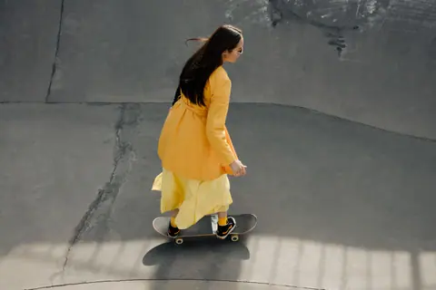 Woman in yellow dress skateboarding in concrete park bowl on a sunny day.