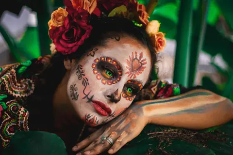 Striking Day of the Dead portrait with colorful makeup and floral headpiece set against lush greenery.