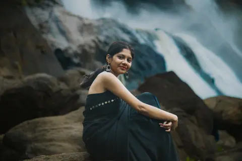 A woman in a black sari seated by rocks near a scenic waterfall in Pariyaram, India.