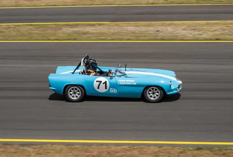 Blue vintage race car with number 71 speeding along a track during a sunny day race.