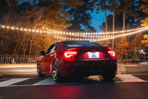 A red sports car parked on an asphalt road with twinkling string lights and a forest backdrop at night.
