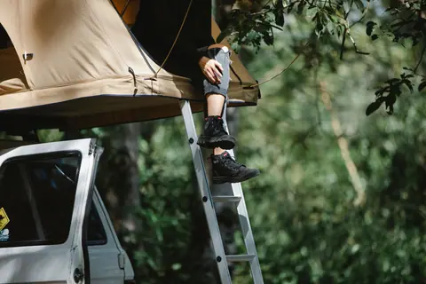 Low angle of crop faceless camper sitting in tent placed on SUV car roof in green forest on sunny day
