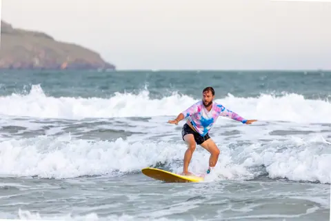 Surfer riding a wave on the ocean with a colorful surfboard and casual attire.
