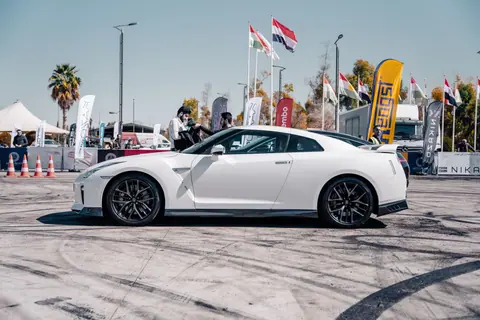 White Nissan GT-R at an outdoor car event, showcasing under blue skies with various flags.