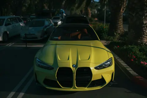 Bright yellow BMW M4 in a shaded parking lot with surrounding greenery, showcasing modern automotive design.