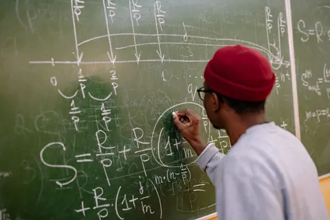 Student writing complex mathematical equations on a chalkboard in a classroom setting.