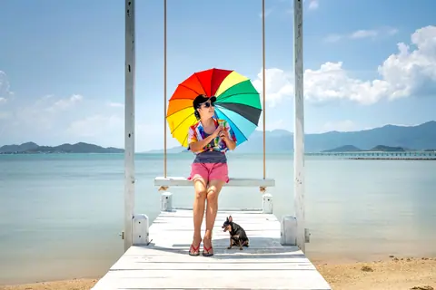 Asian woman with dog enjoys a vibrant beach day on a swing with rainbow umbrella and ocean view.