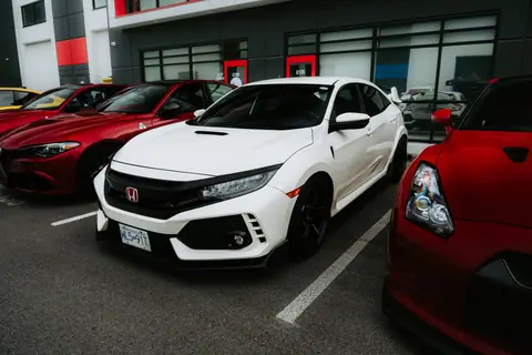 White sports car parked alongside red cars in outdoor lot, modern architecture backdrop.