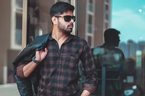 Trendy man with beard wearing sunglasses and plaid shirt, holding a leather jacket outdoors.