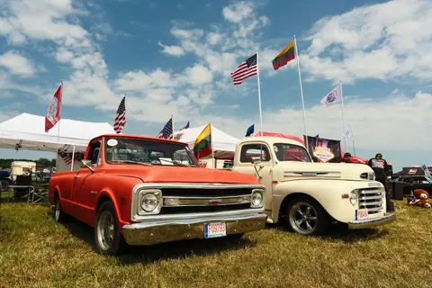 A colorful outdoor exhibit of vintage Chevrolet and Ford trucks amidst flags and tents at a car show.