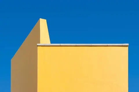 A minimalist architectural shot of a yellow wall contrasted with a clear blue sky.