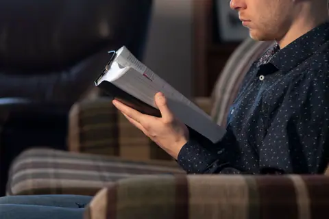 Adult male reading a Bible while seated on a comfortable chair indoors.