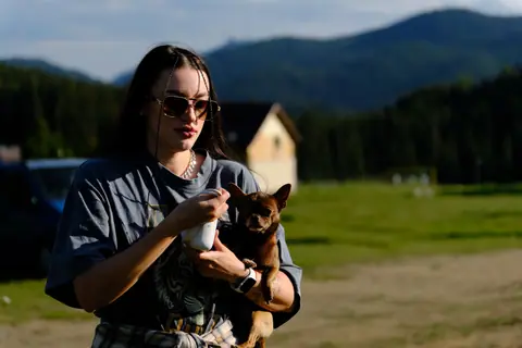 Young woman with sunglasses holding a Chihuahua outdoors against a mountain backdrop in summer.