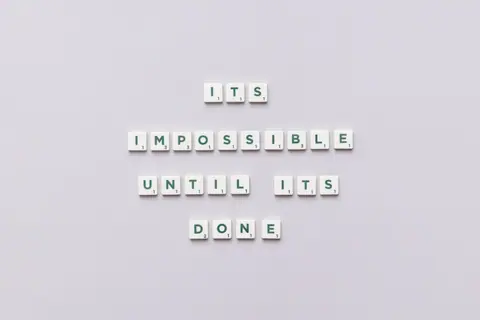 Motivational phrase 'It's impossible until it's done' arranged with Scrabble tiles on a gray background.
