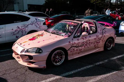 A pink customized car with cherry blossom art displayed at an outdoor car show event.