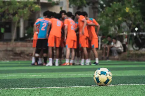 A soccer ball on the pitch with a teenage team strategizing in the background.