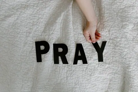 A child's hand placing black letters spelling 'PRAY' on a white quilted blanket.