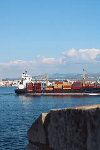 A cargo ship loaded with containers sails past a coastal city with clear blue skies.