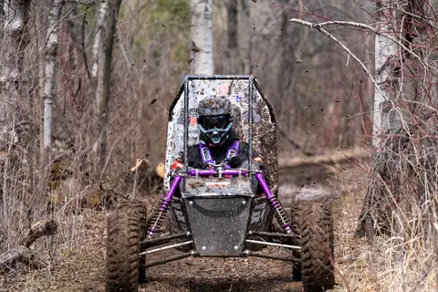 A muddy off-road vehicle with a helmeted driver speeds through a forest trail.