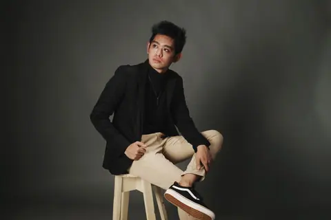 A confident young man poses stylishly in a studio setting, wearing a black jacket and beige pants.