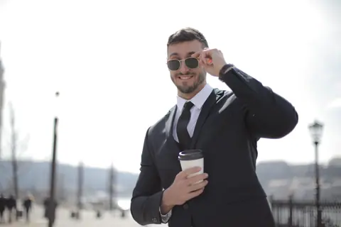 Smiling businessman in suit and sunglasses holding coffee cup outdoors.