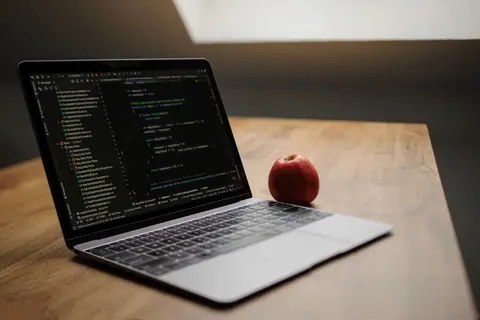 Open laptop displaying code next to a red apple on a wooden desk.