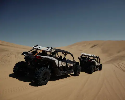 Two off-road vehicles navigate the vast desert sand dunes under a clear blue sky.