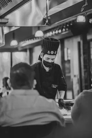 A masked chef serves patrons in an atmospheric black and white restaurant scene, focusing on service.
