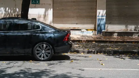 A black sedan parked beside a closed storefront on a sunny day with fallen leaves.