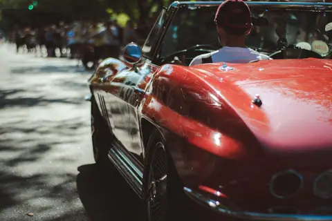 A vintage red convertible driving through a crowd during a sunny summer day.