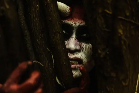 Scary demon-like face with stage makeup peeking through branches in a dark forest.