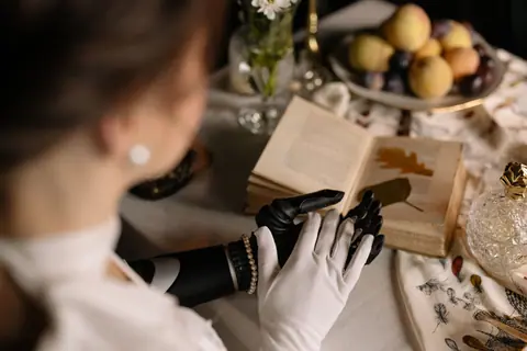 A woman with a prosthetic arm reads a vintage book surrounded by dried leaves and fruit.
