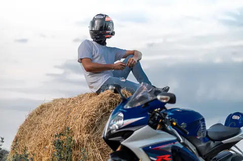 A motorcyclist in a helmet sits on a hay bale beside a blue motorcycle under a cloudy sky.
