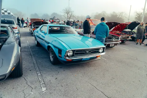 Classic blue Mustang displayed at an outdoor car show with enthusiasts gathered around.