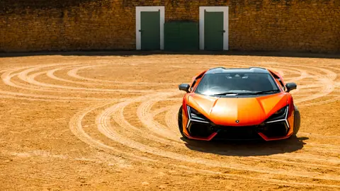 Vibrant orange sports car on dusty track in a sunlit courtyard.