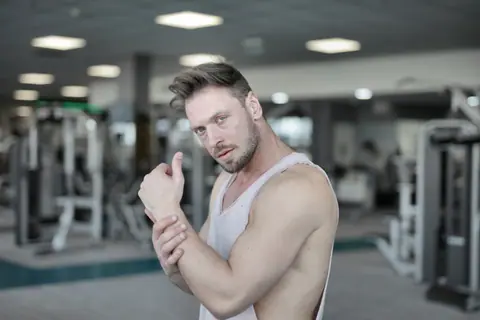 Side view of strong male athlete in casual sportswear standing with raised crossed hands near chest while resting after workout in modern fitness center and looking at camera