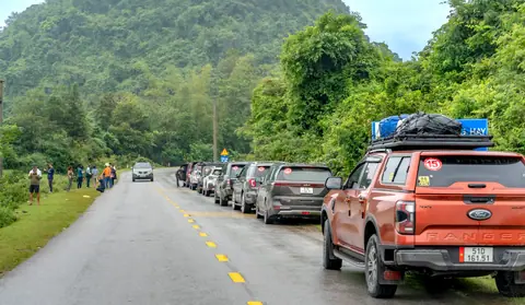 Vehicles parked along a lush green hillside road with people gathering nearby.