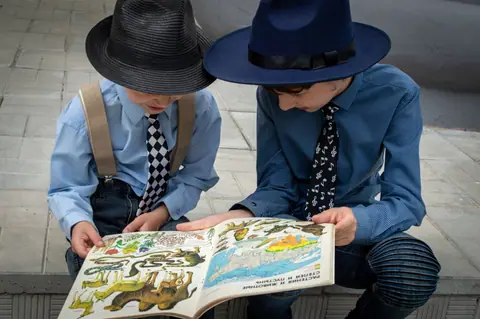 Two young boys wearing fedoras and ties reading an illustrated animal book outside.
