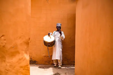 A man in traditional attire plays a drum in a vibrant orange corridor, creating a cultural atmosphere.