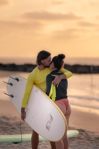 Couple embracing on Tel Aviv beach at sunset holding surfboard, capturing romance and surfing lifestyle.