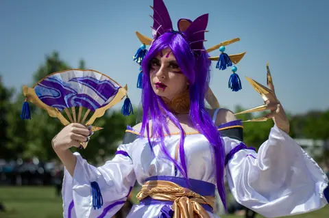 Colorful cosplay character in detailed costume with purple wig and fan outdoors.