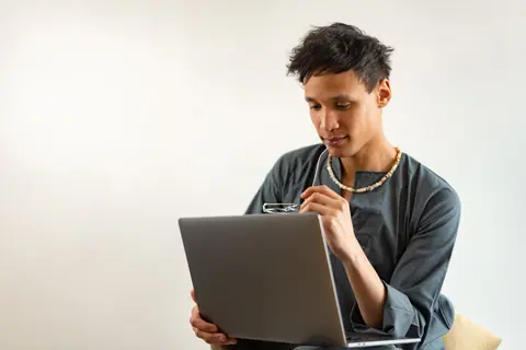 A young man holding eyeglasses, thinking while using a laptop indoors, in a professional setting.