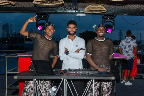 Three men smiling at an outdoor rooftop party with DJ equipment at twilight.