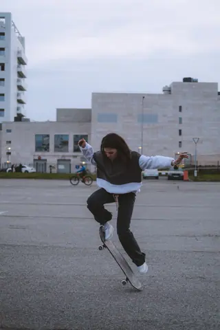 A woman performs an ollie on a skateboard in a city environment, showcasing street fashion and culture.