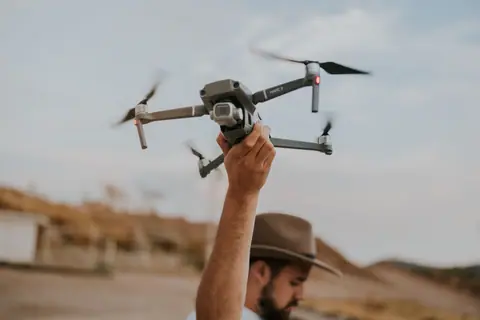 A man in a hat launches a drone in a desert-like outdoor setting during the day.
