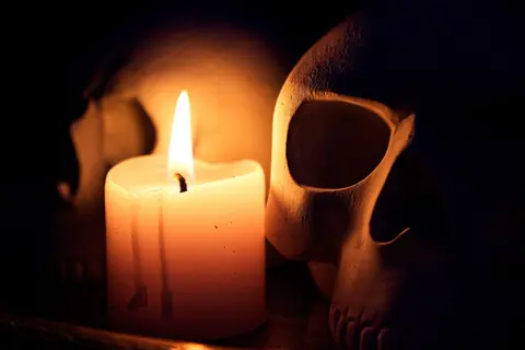 A dramatic still life featuring a candle illuminating two human skulls in the dark.