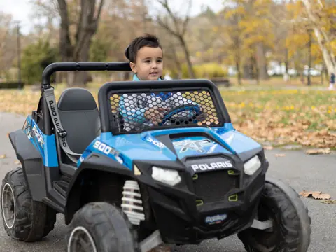 Young boy driving a blue electric toy car outdoors in a park full of autumn leaves.