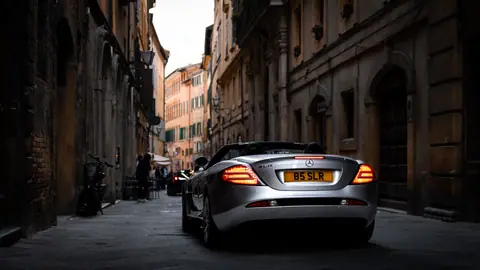 Silver luxury convertible driving through narrow, historic city street at dusk.
