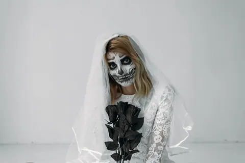 Spooky bride in white dress and skeleton makeup holding black roses. Perfect for Halloween themes.