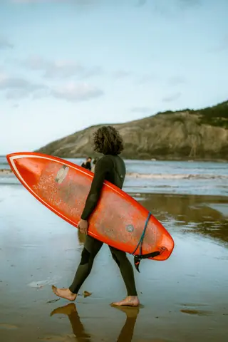 A surfer carrying a red board on a sandy beach with a scenic coastal backdrop.